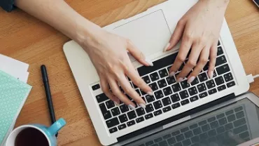 Person typing on a laptop at a wooden desk with glasses, notebook, coffee cup, and plant.